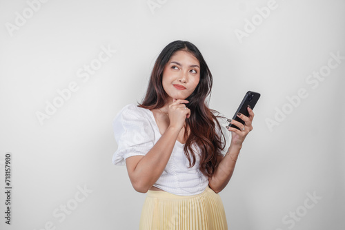 A thoughtful Asian woman is holding her smartphone while imagining her thoughts, isolated by white background.