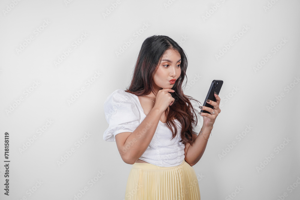Naklejka premium A thoughtful Asian woman is holding her smartphone while imagining her thoughts, isolated by white background.