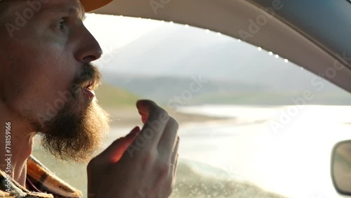 A man with a ginger beard and a cap on his head eats a sweet wafer, putting it in his mouth and chewing it while sitting inside a car overlooking a mountain landscape. Snack during the trip photo