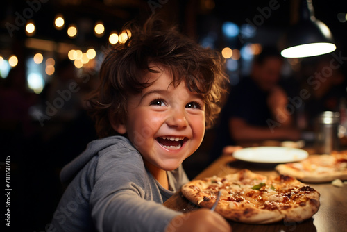 A happy laughing curly-haired boy at the table is eating fresh pizza  holding food in his hands. A joyful atmosphere in the pizzeria