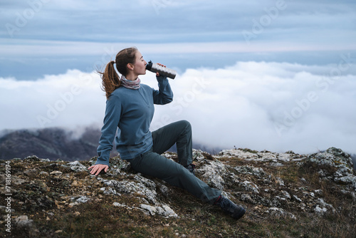 A young beautiful girl with straight brown hair in a ponytail drinks from a thermal glass in nature, tourist resting outdoors, in full view photo
