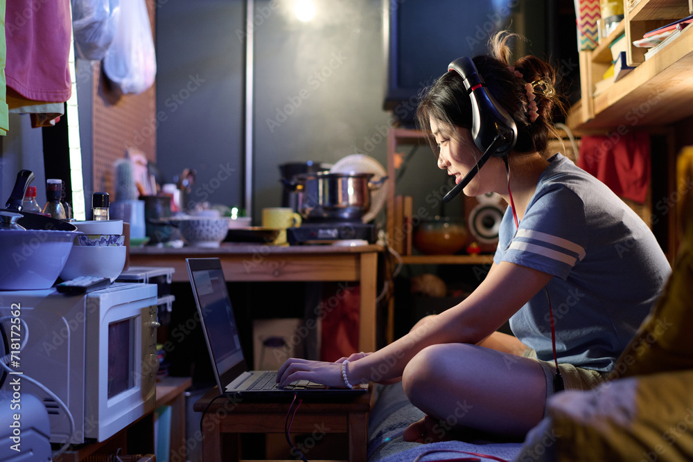 Asian girl in headset speaking in microphone and typing on laptop keyboard while sitting on bed in small apartment