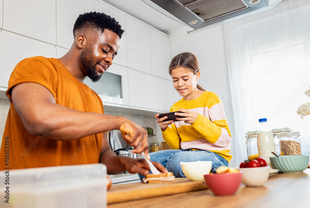 Young single father preparing breakfast in the kitchen at home while his daughter using smart phone. Young single father preparing breakfast in the kitchen at home while his daughter using smart phone.