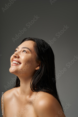 dreamy and young asian woman with brunette hair and acne prone skin smiling on grey background