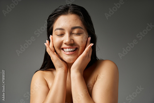 happy and young asian woman with brunette hair and acne prone skin smiling on grey background