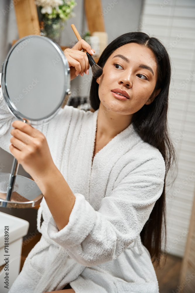 brunette and young asian woman with blemishes applying face powder and looking at mirror, acne