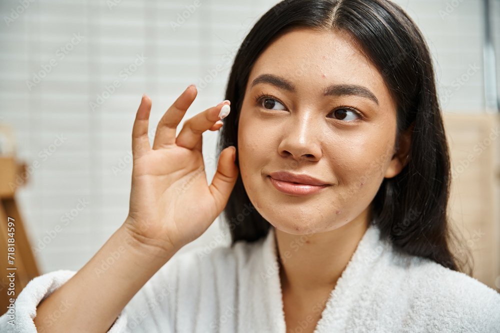 close up of young asian woman with acne applying cream on face and looking at mirror in bathroom close up of young asian woman with acne applying cream on face and looking at mirror in bathroom