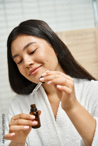 joyful and asian woman in bath robe holding dropper with facial serum to treat acne on face