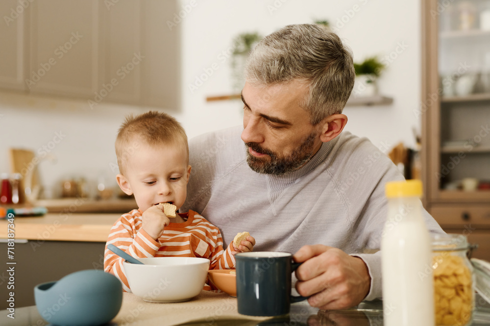 Bearded mature man sitting by kitchen table and looking at his adorable baby son eating small tasty cookies during breakfast Bearded mature man sitting by kitchen table and looking at his adorable baby son eating small tasty cookies during breakfast
