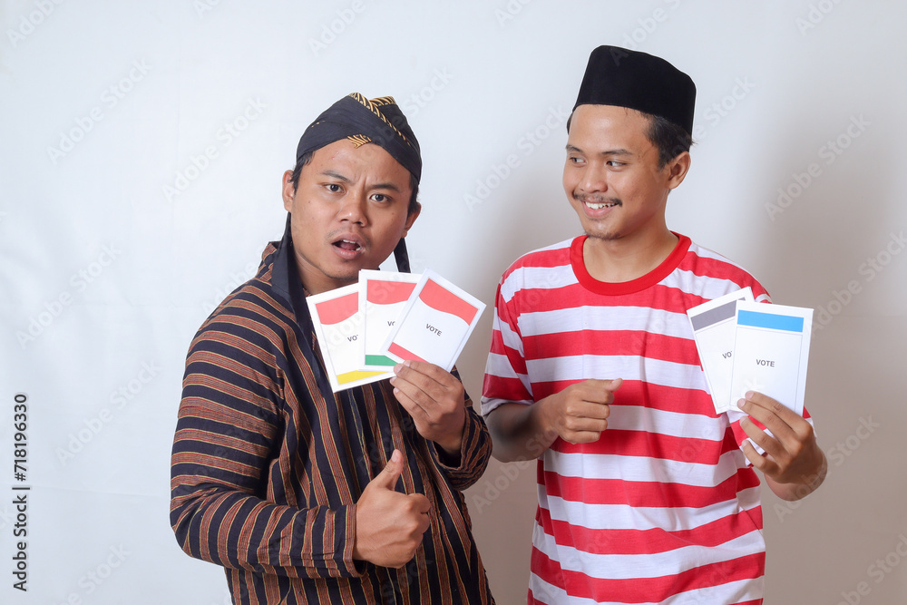 Portrait of two excited Indonesian man from Javanese and Madurese tribes holding voting paper for General Election (Pemilu) of president and government of Indonesia. Isolated image on gray background