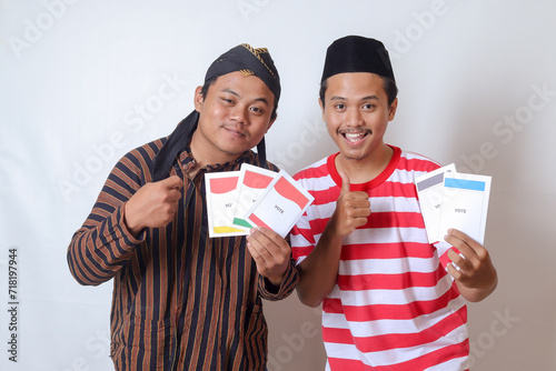 Portrait of two excited Indonesian man from Javanese and Madurese tribes holding voting paper for General Election (Pemilu) of president and government of Indonesia. Isolated image on gray background