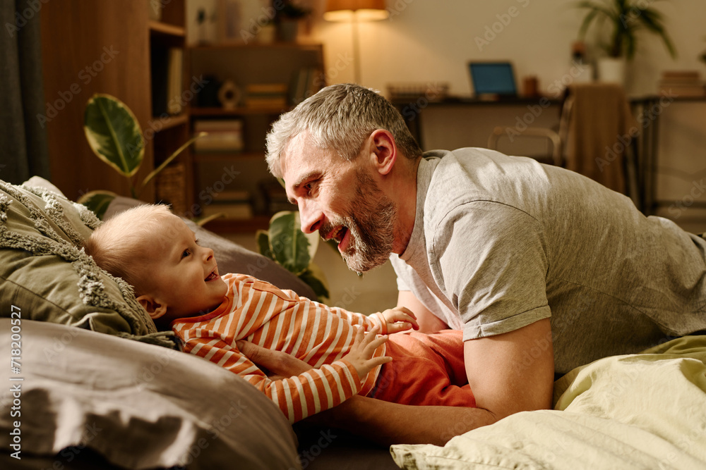 Side view of happy mature man with grey hair and beard playing with his sweet baby son on bed or couch in living room