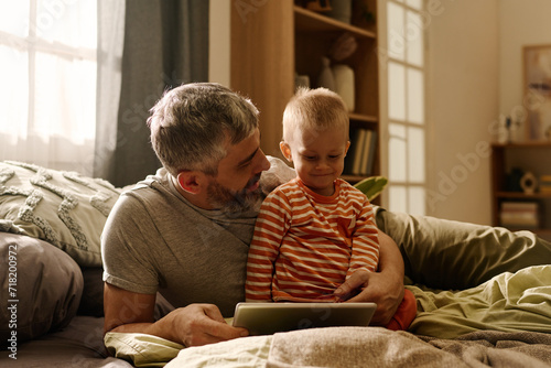 Mature father with tablet looking at smiling baby son while both relaxing on bed or couch in front of camera and watching online cartoon