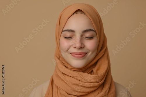 happy young Muslim female in hijab smiling eyes closed while standing against beige background