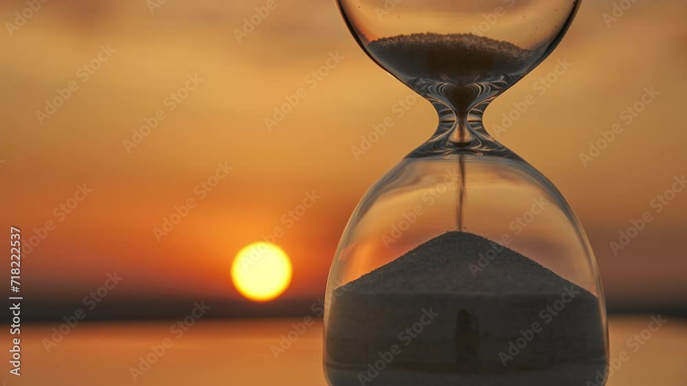 Time concept Time lapse hourglass on blurred background of golden sunset sun disk over horizon sky in sunset rays in summer closeup. Golden sand pours quickly