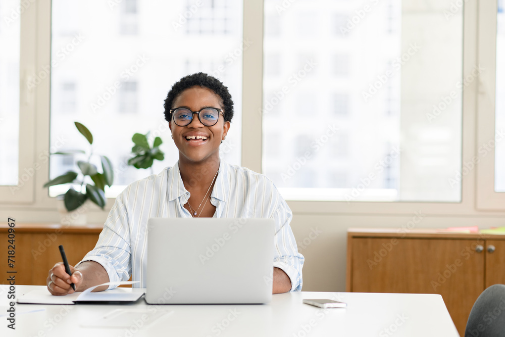 Overjoyed african female student, office worker using laptop for web searching, surfing. Purposeful woman in casual shirt making notes in notebook, making researching, looking at camera and laughing Overjoyed african female student, office worker using laptop for web searching, surfing. Purposeful woman in casual shirt making notes in notebook, making researching, looking at camera and laughing