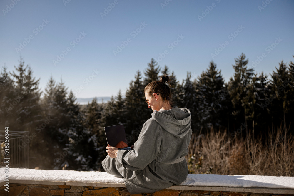 Fototapeta premium Frau im Bademantel sitzt auf einer verschneiten Steinmauer in der Natur und arbeitet am laptop