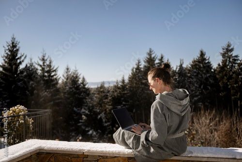 Frau im Bademantel sitzt auf einer verschneiten Steinmauer in der Natur und arbeitet am laptop