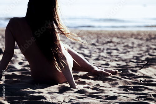 Rear view of lovely naked naturist lady sits on nudist public sea beach, resting. Nude perfect woman with sexy body, from behind. Nudism naturism lifestyle concept, clothing optional. Copy text space