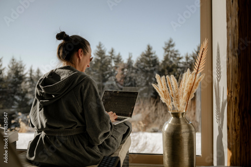 Frau im Bademantel sitzt mit einem Laptop am Schoß in einem WIntergarten