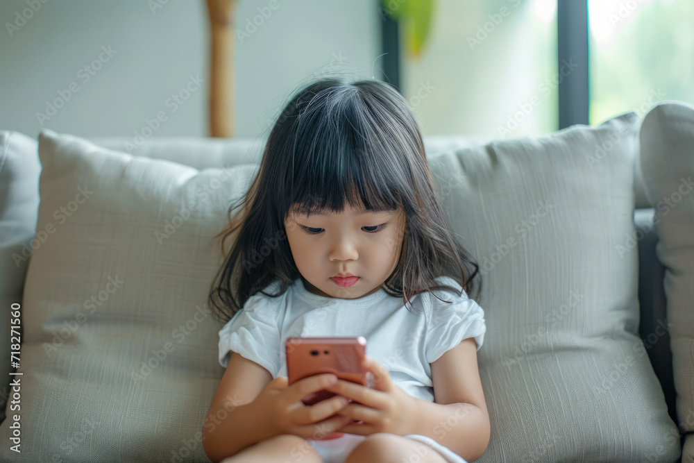Asian child sitting on a sofa, browsing internet on mobile device and playing with smartphone apps Asian child sitting on a sofa, browsing internet on mobile device and playing with smartphone apps