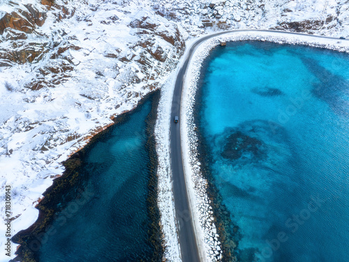 Fototapeta Naklejka Na Ścianę i Meble -  Aerial view of snowy road to the island and blue sea on both side. Bridge on snow and azure transparent water in winter. Landscape. Top drone view of road to the Henningsvaer, Lofoten islands, Norway