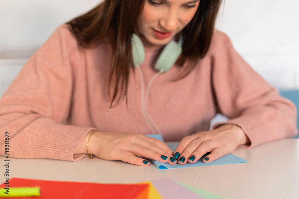 Fototapeta premium Woman in Pink Sweater with Headphones, Folding a blue paper, preparing a playful April Fools' surprise.