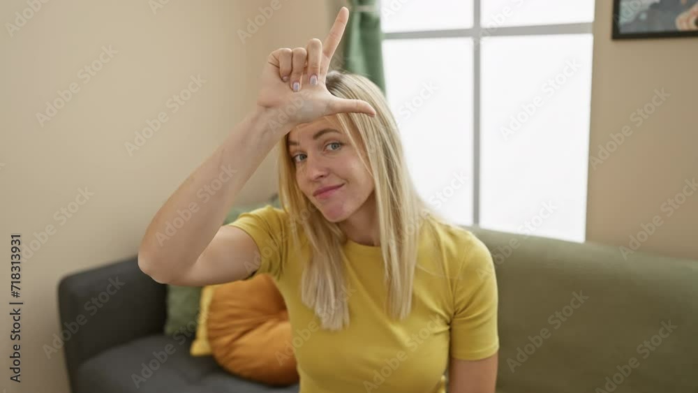 At home, young blonde woman wearing a t-shirt humorously insults, teasing and making fun of others with a loser symbol gesture. forehead fingers mock in playful rudeness.