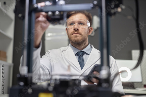 Low angle view of bearded Caucasian man technician in protective glasses adjusting 3D printer in engineering laboratory, look through device frame in blurred foreground