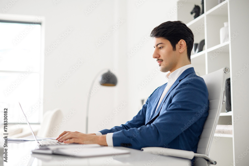 Professional Hispanic businessman in blue suit types attentively on a laptop in a bright modern office, showcasing productivity and digital proficiency, sitting in the office space, side view
