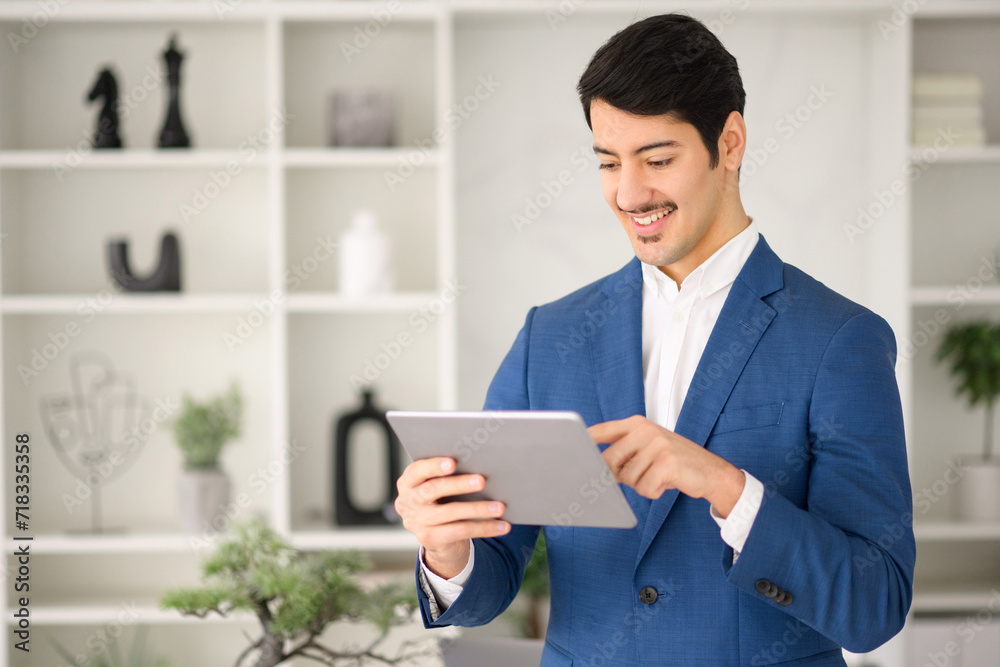Cheerful Hispanic businessman reviews content on digital tablet, his expression one of focus and interest, against a background of a stylish office shelf. Technology in everyday business operations