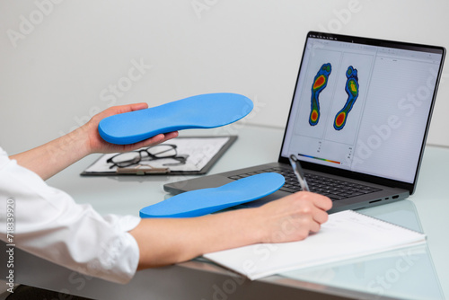 Female doctor orthopedist  holding custom made insoles in a clinic in front of the laptop with a test feet picture.