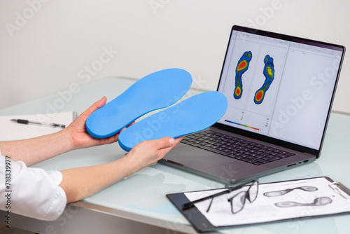Female doctor orthopedist  holding custom made insoles in a clinic in front of the laptop with a test feet picture.  Feet recreation and orthotic medicine concept