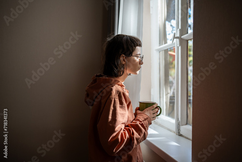 Lonely sad woman with cup of tea looking a window. Unsociable teenage girl spending weekend at home because of friendless, social difficulties, problems in communication, misunderstanding