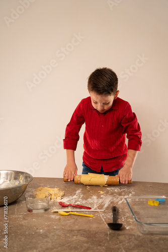 Niño chef amasando y haciendo galletas para San Valentín aplanando la masa con rodillo y utilizando utensilios y recipientes para hornear y cocinar photo