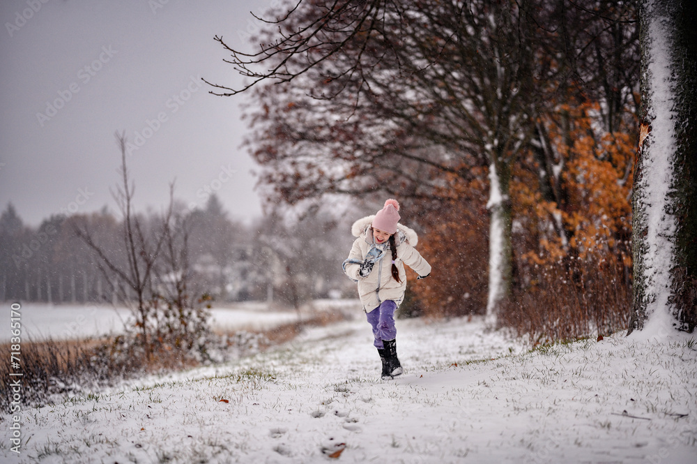 a small child in winter clothing joyfully running on a snowy path lined with bare trees and autumn leaves