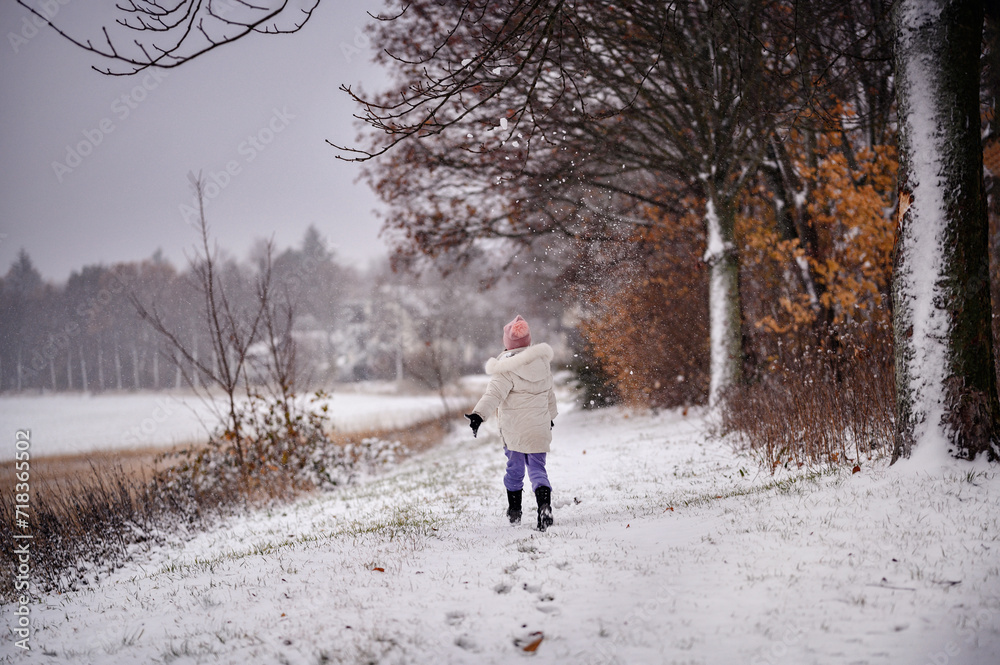 The photo captures a child in a white coat and purple pants joyfully running in a snowy landscape, with snowflakes gently falling around.