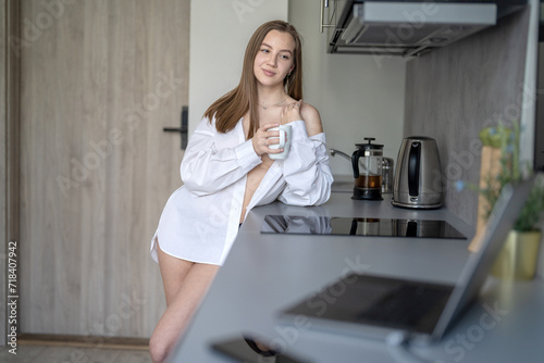 Beautiful Young Girl is Standing in the Kitchen and Drinking Coffee. Morning Routine. Wearing White shirts. Blurry Laptop and Mobile Phone on the Desk.