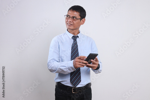 Adult Asian executive man with glasses and formal clothes holding smarpthone while looking away thinking