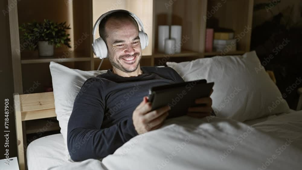 Smiling bearded man wearing headphones using tablet in bed at night Smiling bearded man wearing headphones using tablet in bed at night