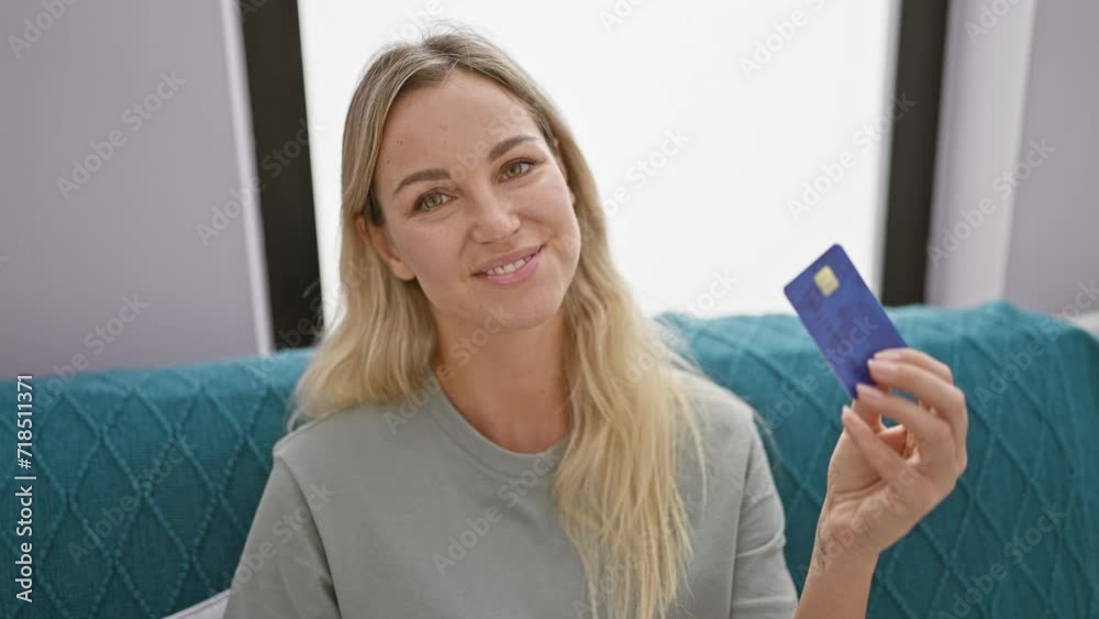 A cheerful, blonde woman holds a bank card, comfortably posing in a modern living room, embodying financial independence. A cheerful, blonde woman holds a bank card, comfortably posing in a modern living room, embodying financial independence.