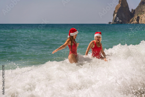 Women Santa hats ocean play. Seaside, beach daytime, enjoying beach fun. Two women in red swimsuits and Santa hats are enjoying themselves in the ocean waves. © svetograph