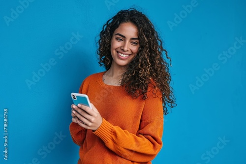 Young adult smiling happy pretty latin woman holding mobile phone device, doing ecommerce online shopping on cell, using apps on cellphone standing isolated on blue, Generative AI photo