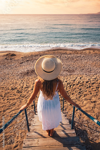 Relaxing young woman in white dress and hat enjoying sunset on the beach
