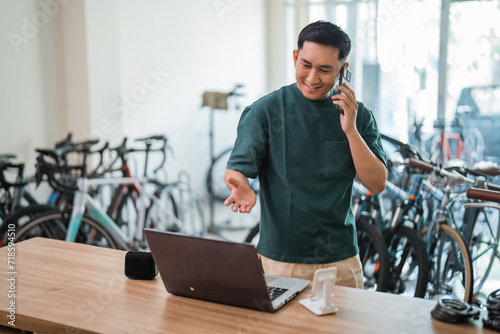 young man makes a cell phone call while working on a laptop at a desk inside a bicycle shop