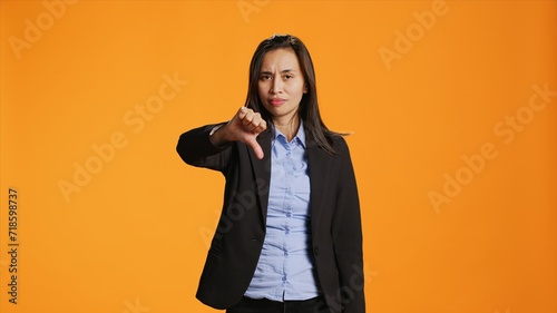 Asian female model giving dislike in front of camera, showing thumbs down with displeased facial expressions. Businesswoman does rejection and disapproval gesture over backdrop.