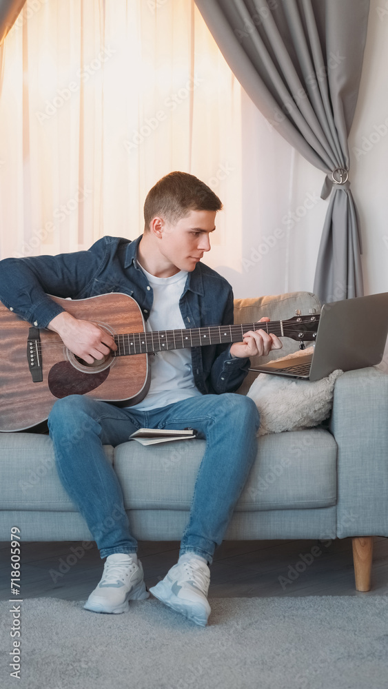 Guitar practice. Online music course. Talented focused man playing song chords on string instrument at remote laptop lesson on couch in living room at home.