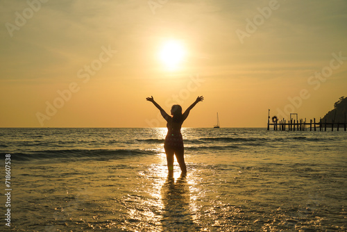 Asian women in bikini swimsuit silhouette with raised arms against calm sunset beach. Happy woman enjoying the summery sunset at the lake on her vacation.