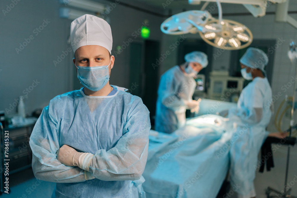 Portrait of male doctor in surgical uniforms and masks standing posing looking at camera with folded arms in operating theater. Diverse professional team of surgeons performing operation on background Portrait of male doctor in surgical uniforms and masks standing posing looking at camera with folded arms in operating theater. Diverse professional team of surgeons performing operation on background