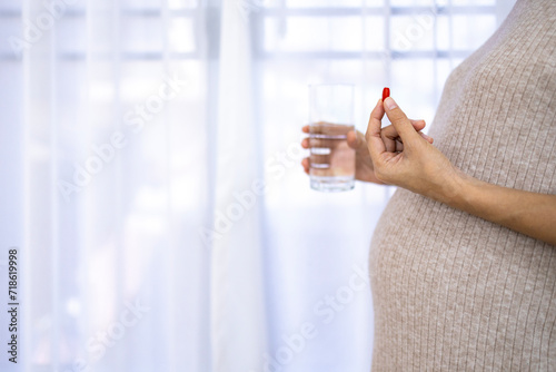 Portrait of pregnant woman standing near window with white curtain.Pregnancy take drug or vitamin with glass at home with copy space.Pregnancy, motherhood,concept.Antenatal care.